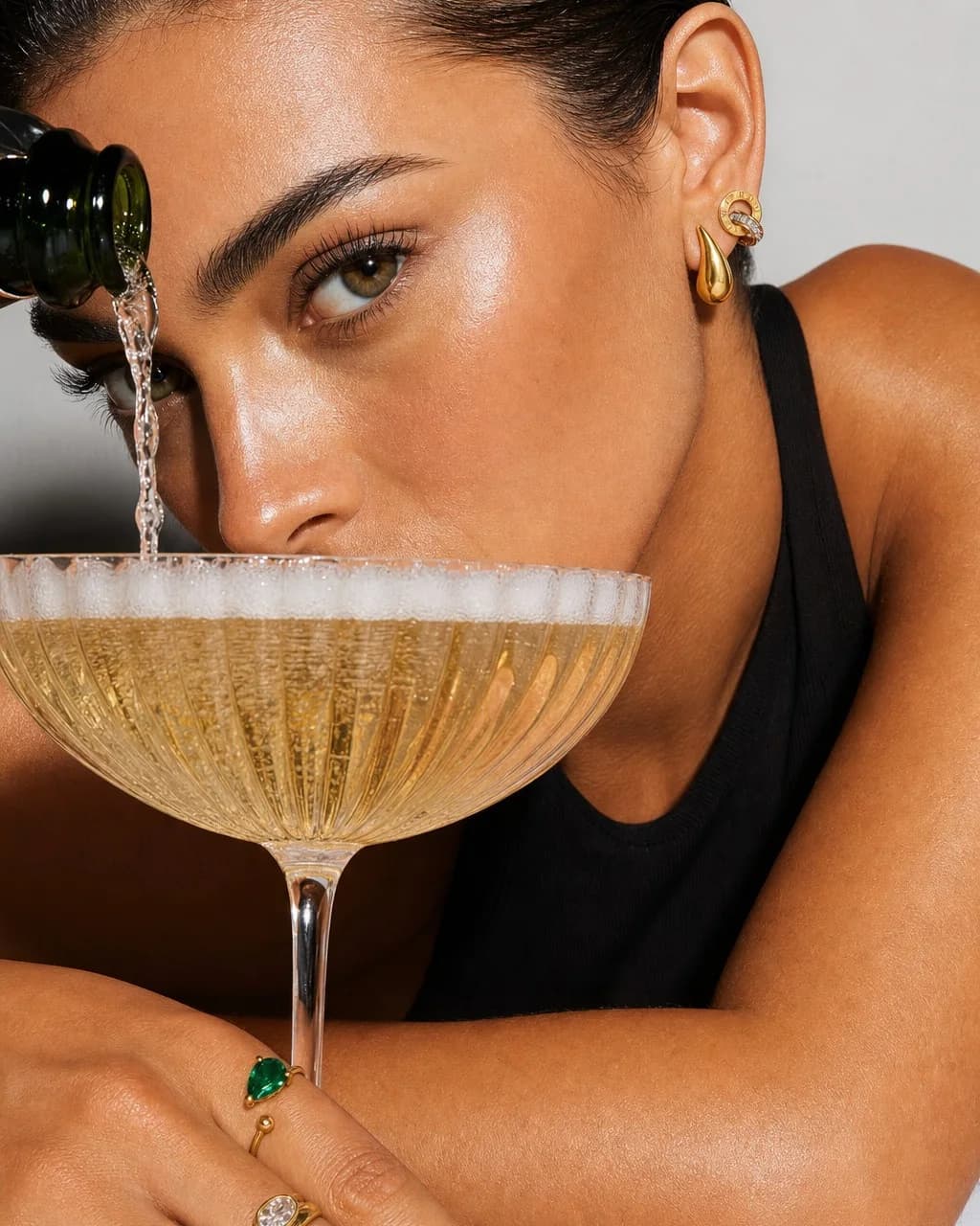 Champagne poured into a coupe at eye level, wearing the Chunky Teardrop Stud and Double Ring Stud, an emerald ring on the hand
